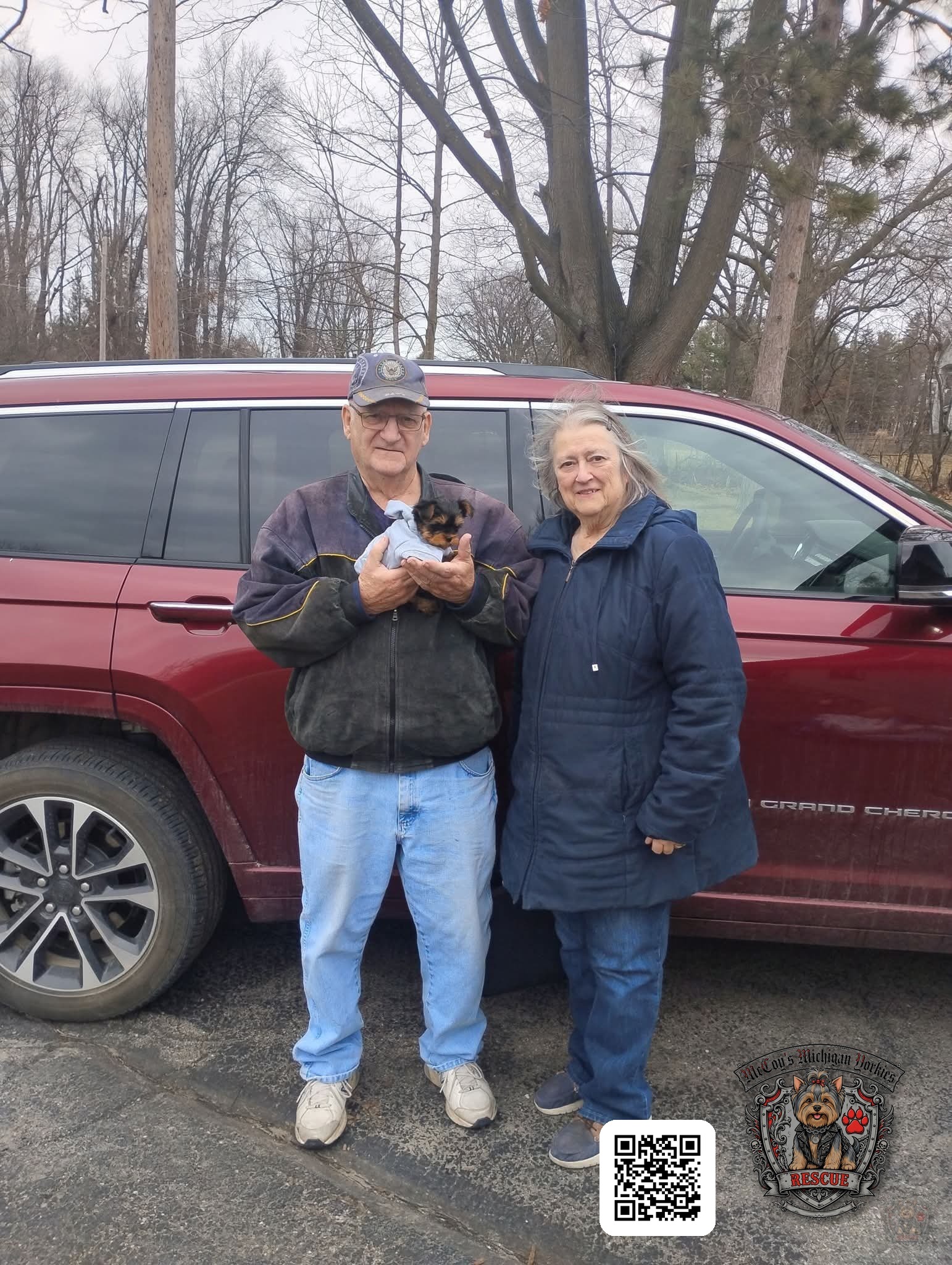 A returning McCoy's family smiling beside their red Jeep while holding their tiny new Yorkshire Terrier puppy